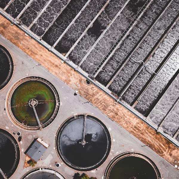Aerial view of a water treatment plant featuring circular clarifier tanks and long, rectangular aeration basins filled with bubbling water, separated by a dirt access road.